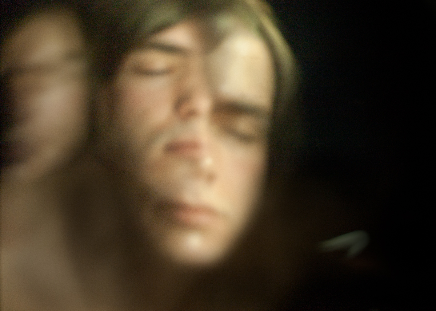 A softly blurred, close-up self-portrait of a person with eyes closed, rendered in warm amber tones, evoking introspection and the early experimental work exploring identity through distorted imagery.