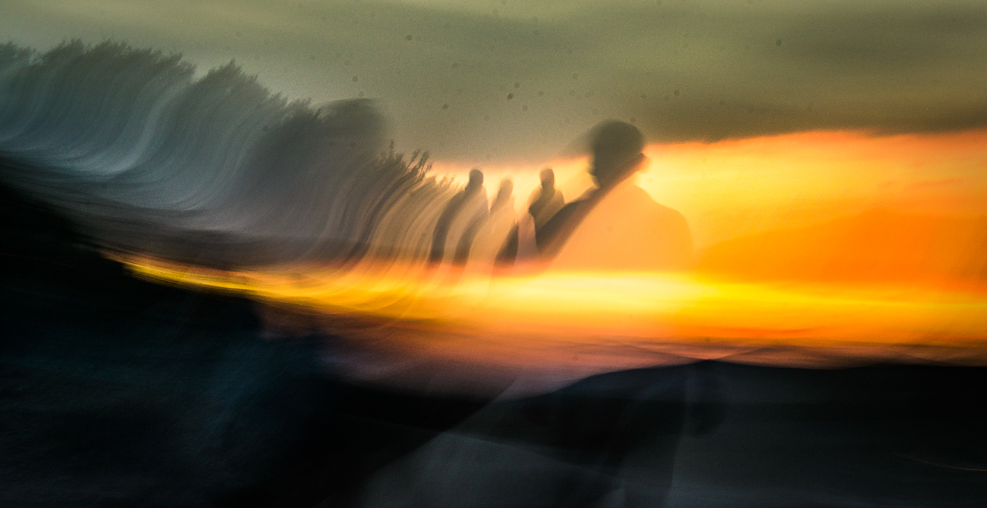 An abstract ICM photograph of silhouetted figures against a blazing orange and yellow sunset, with sweeping motion blur streaking across dark coastal rocks, creating a dreamlike, painterly effect.