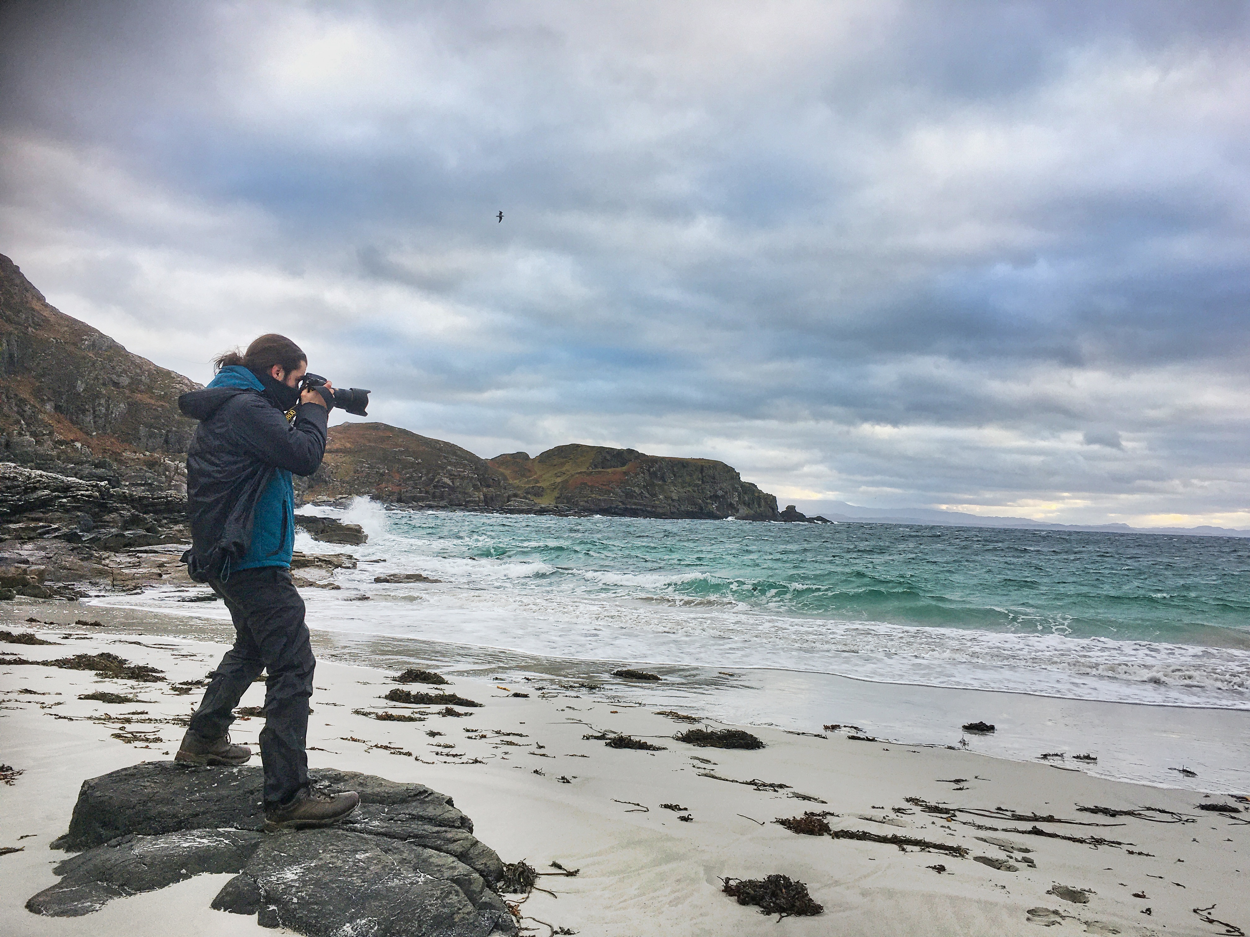 David Julià standing on a rocky white sand beach, camera raised to his eye, facing crashing waves under a dramatic cloudy sky on the Scottish Atlantic coast.