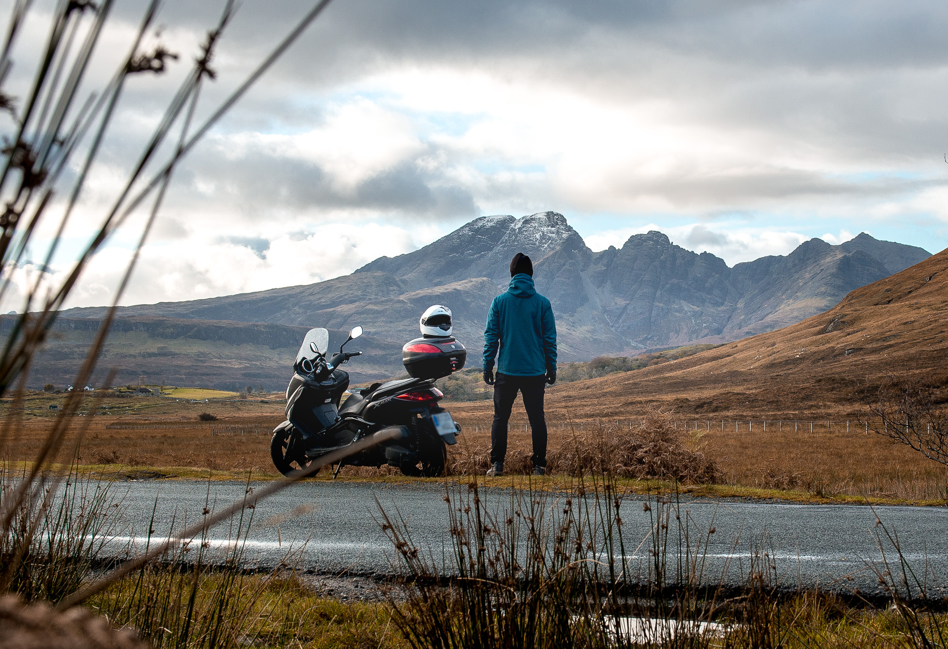A figure in a blue jacket standing beside a parked scooter on a quiet road, looking out toward snow-dusted rugged mountains under an overcast sky, evoking the solitude of a solo winter journey through Scotland.